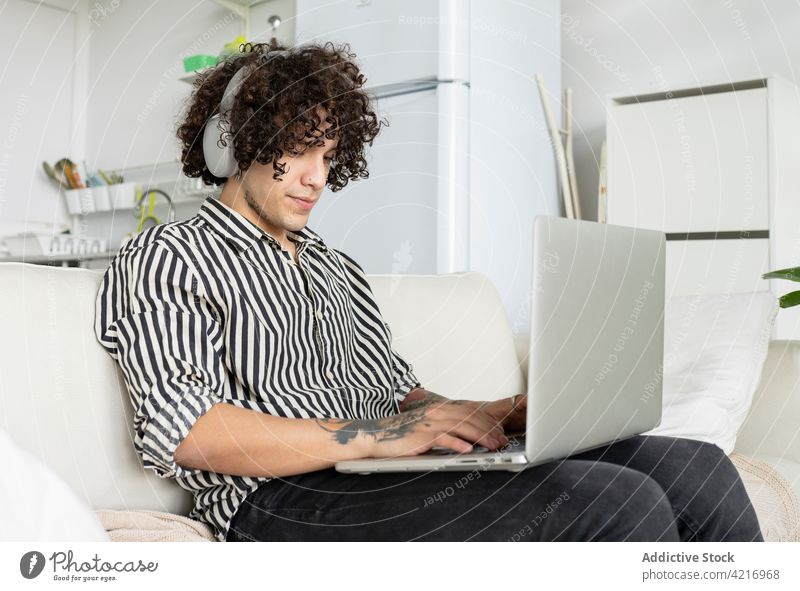 Tätowierter Mann surft im Internet auf einem Laptop auf dem Sofa zu Hause Kopfhörer zuhören online Freizeit Surfen Tattoo heimwärts benutzend Apparatur Gerät