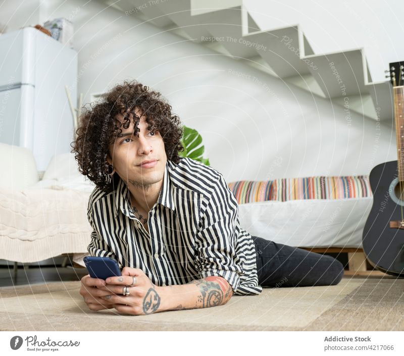 Verträumter Mann mit Smartphone auf dem Boden im Haus liegend verträumt Tattoo nachdenken freundlich herzlich freie Zeit Stock Porträt Gitarre akustisch Musik