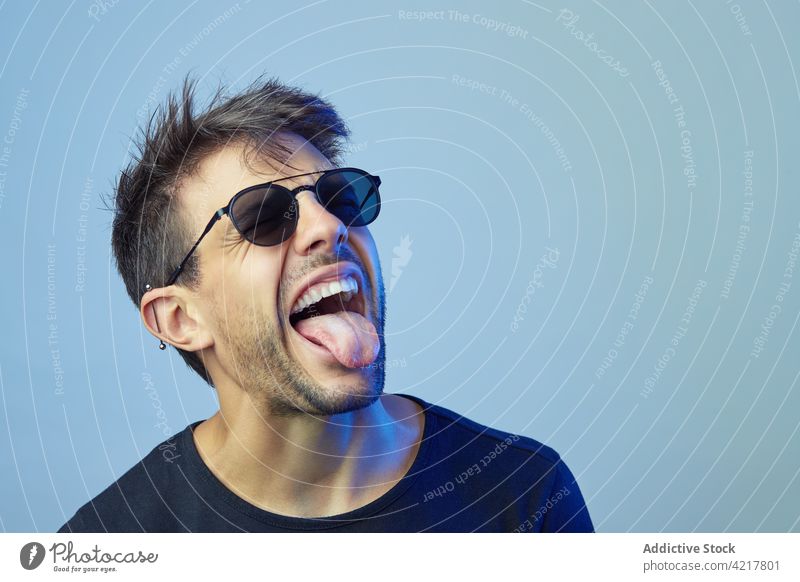 Provokativer Mann zeigt Zunge im Studio Zunge zeigen ausspannen expressiv auflehnen Grimasse Gesicht machen exzentrisch neonfarbig Licht männlich Sonnenbrille