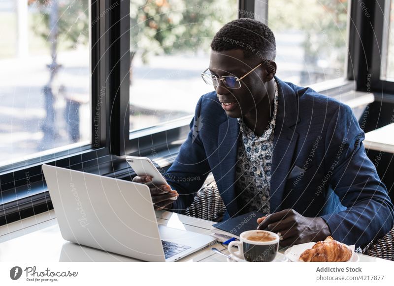 Stilvolle ethnische Führungskraft mit Smartphone und Laptop in einem Cafe Exekutive Arbeit plaudernd gut gekleidet Frühstück Mann Café benutzend Apparatur Gerät