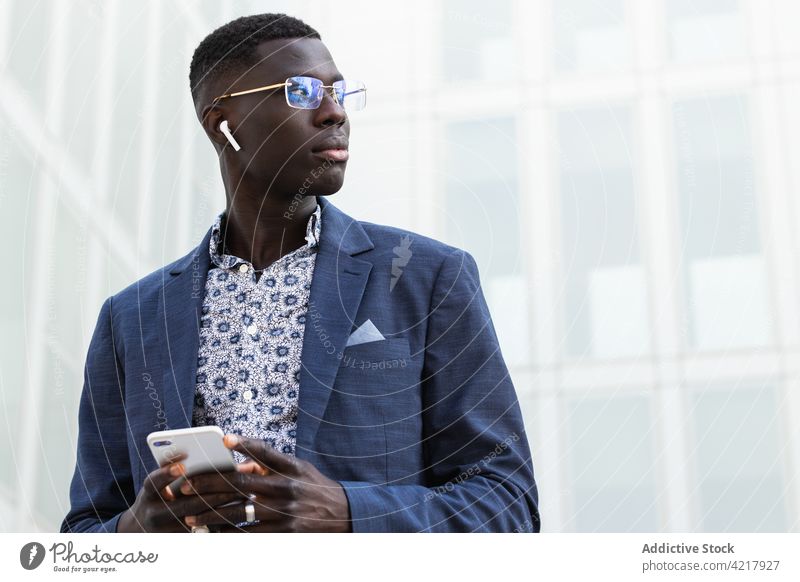 Stilvoller schwarzer Unternehmer im Ohrhörer mit Smartphone Geschäftsmann Ohrstöpsel gut gekleidet selbständig maskulin nachdenken Multimedia Porträt benutzend
