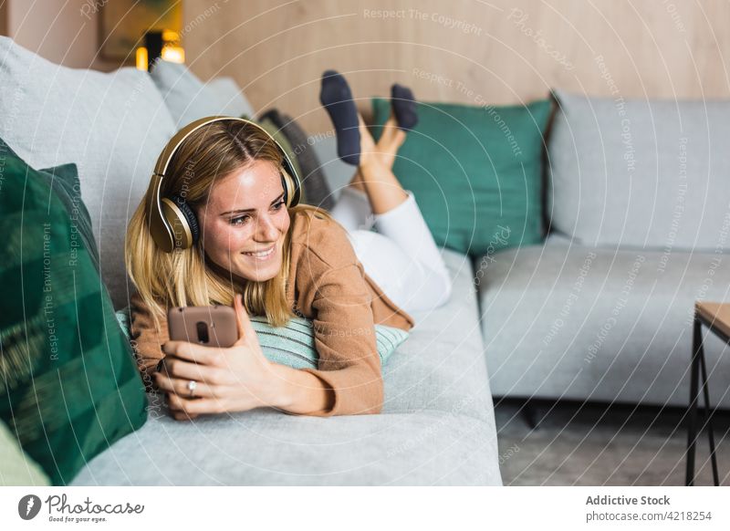 Fröhliche Frau, die mit Kopfhörern auf einem Mobiltelefon Musik hört Zwinkern zuhören heiter heimwärts soziale Netzwerke Gesang genießen Browsen Glück