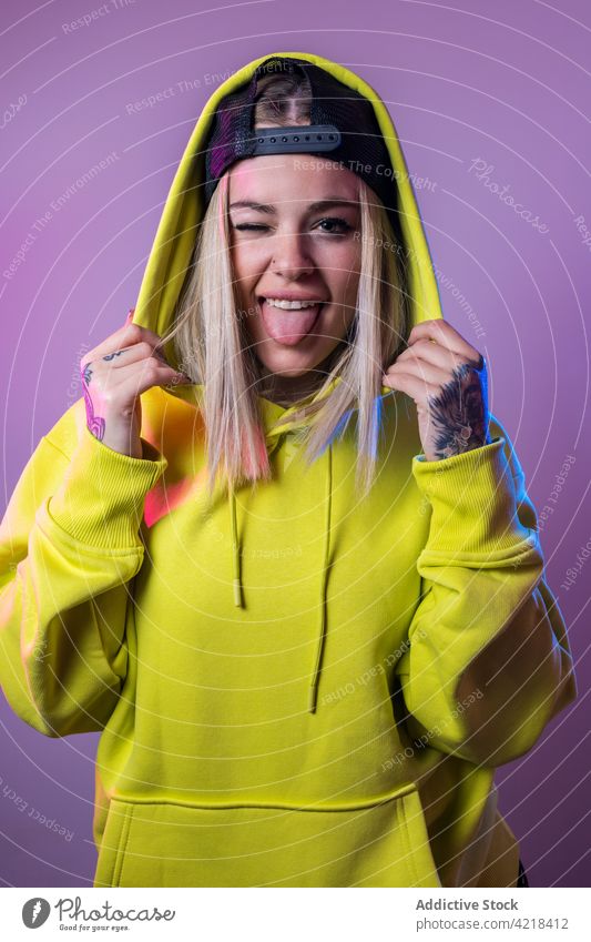 Fröhliche Frau im Kapuzenpulli zeigt Zunge im Studio Zunge zeigen Hipster auflehnen ausspannen Gesichtsausdruck Gesicht machen Grimasse neonfarbig Licht Atelier