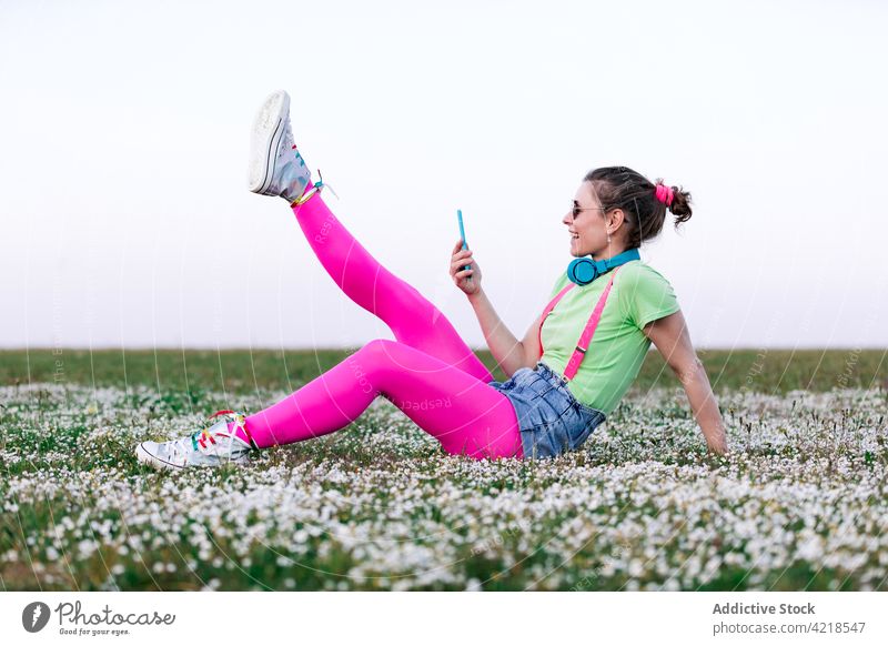 Frau benutzt Smartphone und sitzt mit hochgelegten Beinen auf einer Wiese benutzend Rasen angehobene Beine Landschaft freudig sorgenfrei Apparatur Outfit