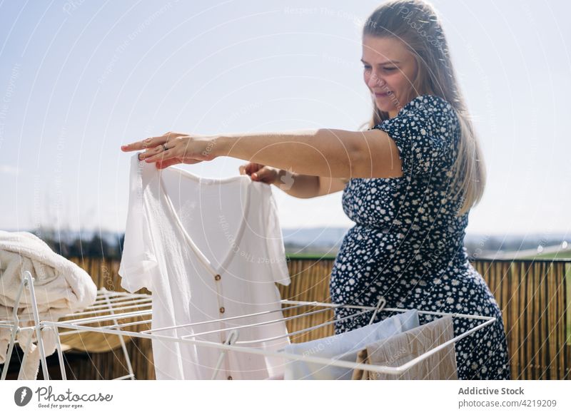 Lächelnde schwangere Frau hängt Wäsche auf einem sonnigen Balkon auf Leinen Wäscherei Trocknung Ablage warten Mutterschaft vorwegnehmen Himmel Schwangerschaft