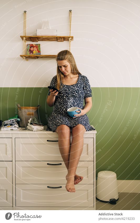 Lächelnde schwangere Frau mit Tee beim Surfen auf dem Smartphone zu Hause Browsen zuschauend Schwangerschaft herzlich pränatal heimwärts benutzend Apparatur