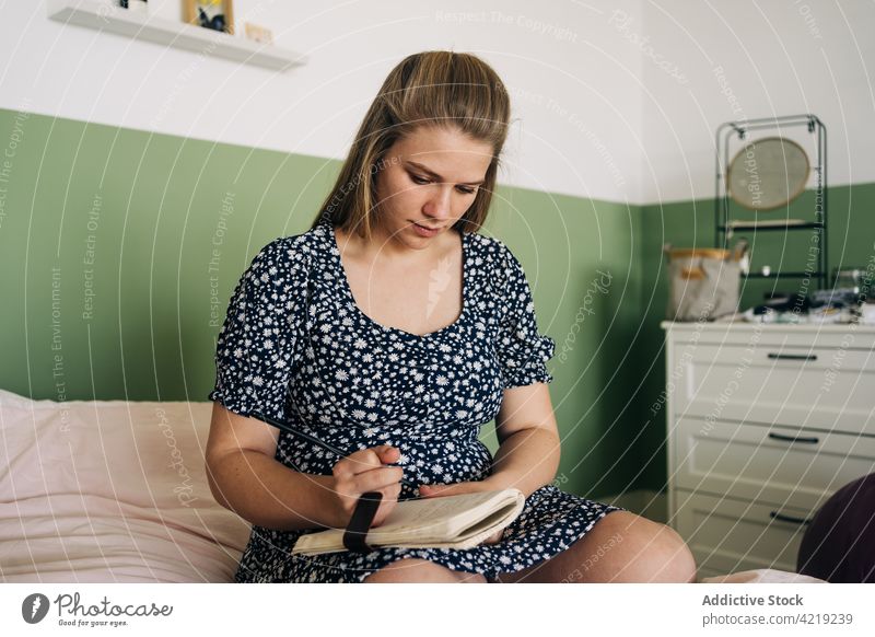 Schwangere Frau sitzt auf dem Bett und schreibt Notizen sitzen zur Kenntnis nehmen Notebook achtsam feminin sanft Haus Schreibstift schreiben Papier Notizblock