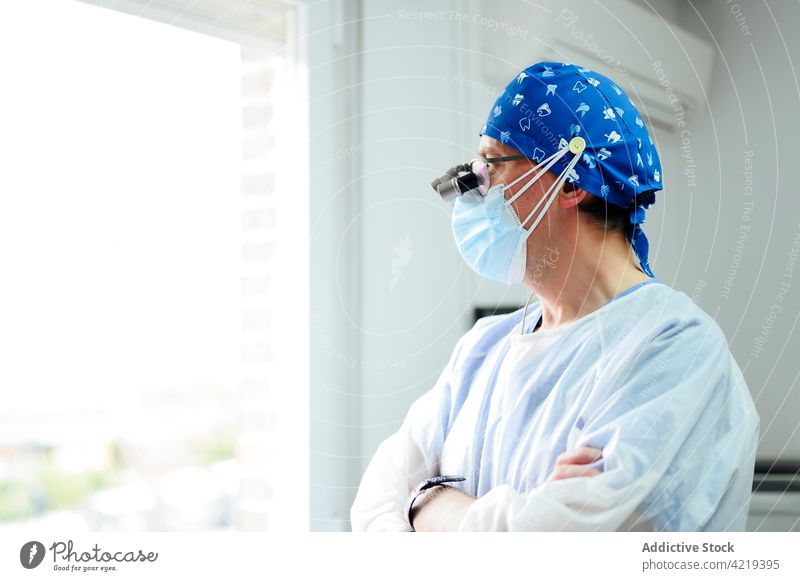 Unbekannter Chirurg mit medizinischem Fernglas und verschränkten Armen in einer Klinik herausschauen Fenster die Arme verschränkt Uniform professionell