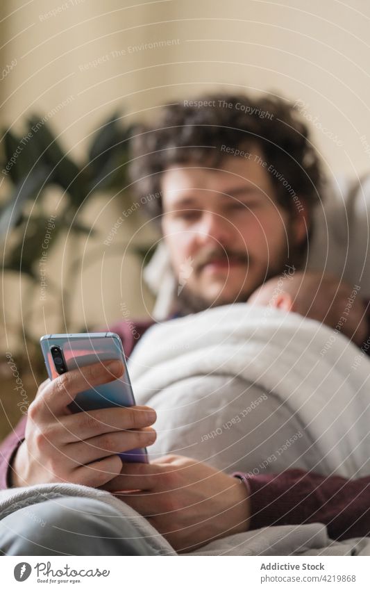 Vater mit unerkennbarem Neugeborenen chattet zu Hause auf dem Smartphone Papa Baby plaudernd freie Zeit Umarmen Vaterschaft Internet heimwärts benutzend