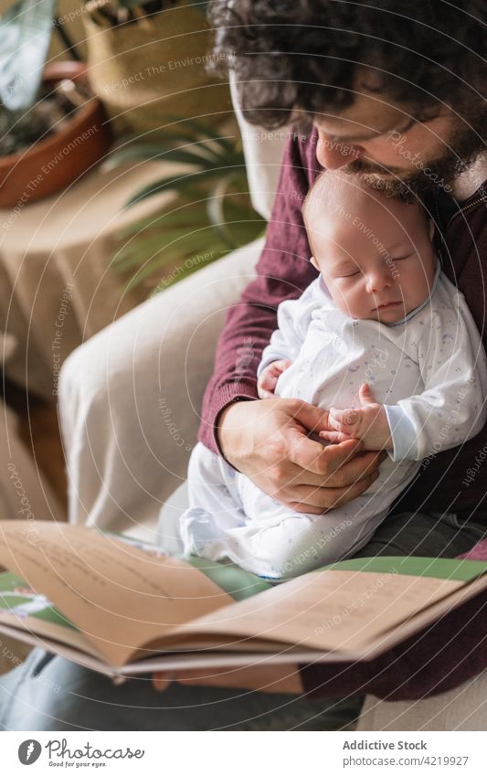 Vater mit neugeborenem Baby liest zu Hause ein Buch Papa lesen Vaterschaft Literatur Zeit verbringen Armsessel Mann Säugling Säuglingsalter charmant idyllisch