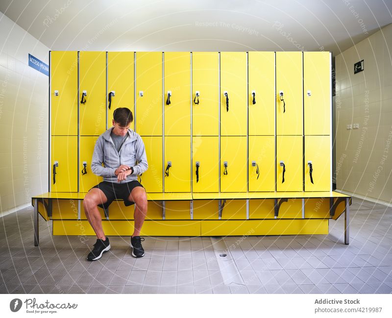 Sportler, der in der Umkleidekabine die Zeit auf seiner Armbanduhr beobachtet prüfen punktuell Bank Spind Raum Stil Mann modern Turnschuh Sportbekleidung sitzen