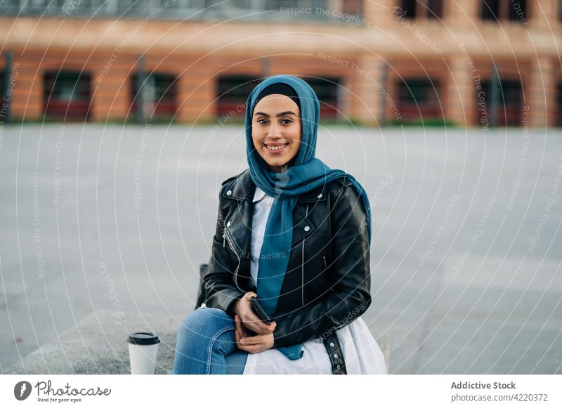 Stilvolle ethnische Frau im Hidschab hält Smartphone auf der Straße Hijab benutzend Surfen Großstadt Bank muslimisch Imbissbude trinken zum Mitnehmen Kaffee
