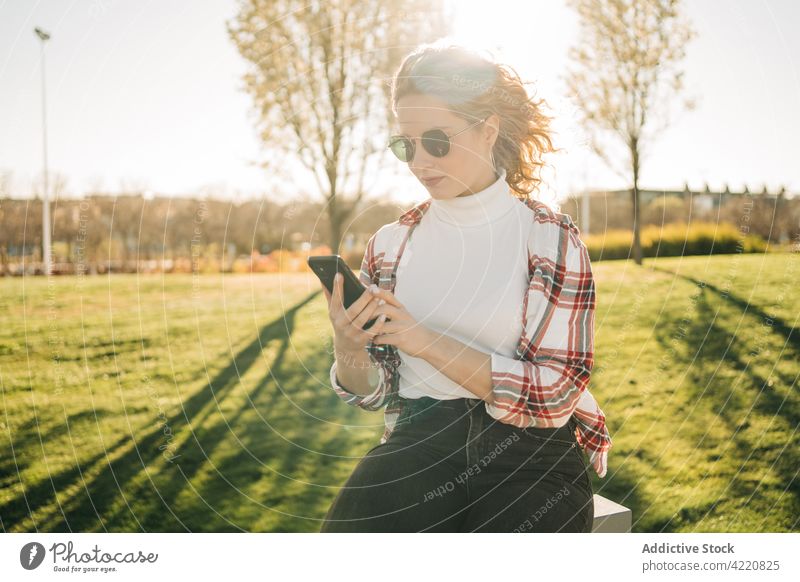 Stilvolle Frau beim Stöbern im Park Smartphone Apparatur Gerät Mobile benutzend modern trendy Telefon krause Haare zuschauend sitzen Bank Sonnenbrille