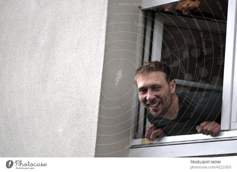 Der freundlichste Nachbar aller Zeiten - ein junger Mann und Vater lehnt sich lachend aus einem kleinen Fenster heraus und schaut direkt in die spontan gezückte Kamera des Fotografierers