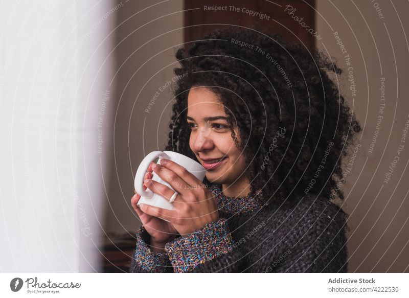 Ethnische Frau trinkt zu Hause ein Getränk trinken Tasse heimwärts Fenster Lächeln Afro-Look Frisur krause Haare ethnisch charmant verträumt genießen
