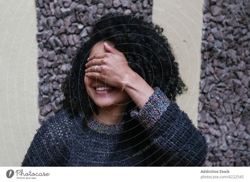 Glückliche ethnische Frau mit lockigem Haar, die ihre Augen mit der Hand bedeckt Afro-Look Frisur Lächeln charmant krause Haare Vorschein Schönheit natürlich