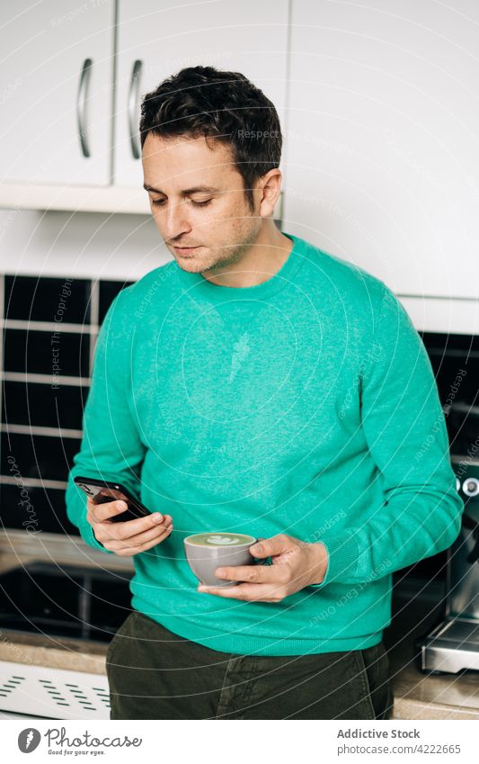 Mann mit Milchkaffee, der in der Küche mit seinem Smartphone chattet plaudernd Latte Kaffee freie Zeit Internet online benutzend Apparatur Gerät Funktelefon