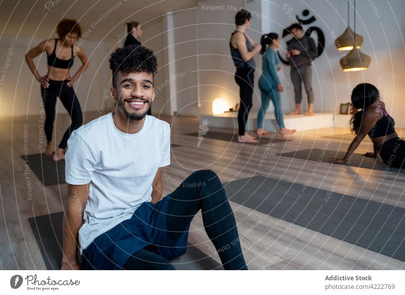 Fröhlicher schwarzer Mann sitzt auf einer Matte im Yogastudio Klasse Atelier Menschengruppe Lektion Lächeln üben Zusammensein heiter rassenübergreifend