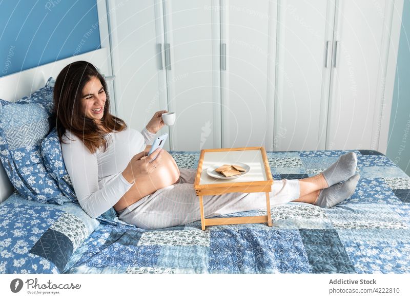 Glückliche schwangere Frau beim Frühstück und beim Surfen auf dem Smartphone Bett Morgen Schwangerschaft Browsen zuschauend Telefonanruf heimwärts Gerät heiter