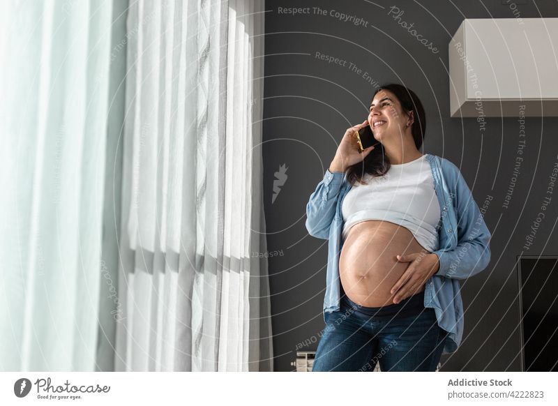 Schwangere Frau, die zu Hause mit ihrem Smartphone spricht schwanger reden sprechen berühren Bauch soziale Netzwerke Schwangerschaft stehen Raum heimwärts