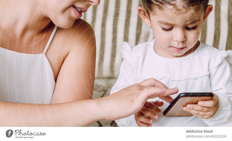 Crop-Mutter und kleines Kind benutzen gemeinsam ein Smartphone Tochter Hilfsbereitschaft benutzend Browsen wenig erklären Zusammensein ethnisch Sofa heimwärts