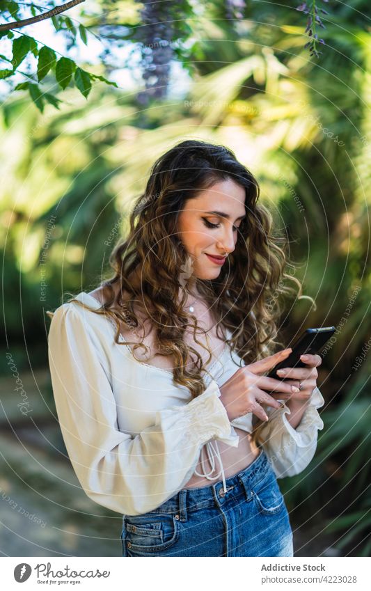 Lächelnde junge Frau benutzt ihr Smartphone in einem üppigen Park benutzend Browsen Zahnfarbenes Lächeln Garten Inhalt Stil sorgenfrei Mobile attraktiv schön