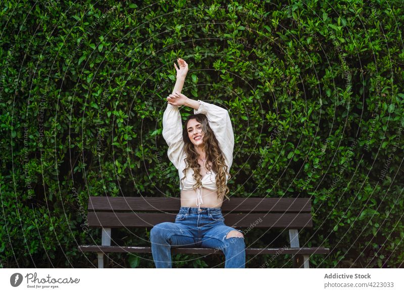 Glückliche junge Frau sitzt mit erhobenen Armen auf einer Parkbank heiter Zahnfarbenes Lächeln Arme hochgezogen traumhaft pflanzlich Anmut Körperhaltung Bank