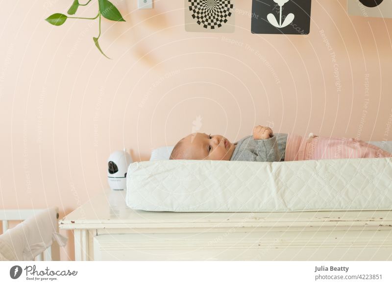 Junges Baby liegt auf einer Wickelauflage in einem pfirsichfarbenen Kinderzimmer; schwarz-weißes Mobile und Babyphone in der Nähe Säugling 3 Monate alt