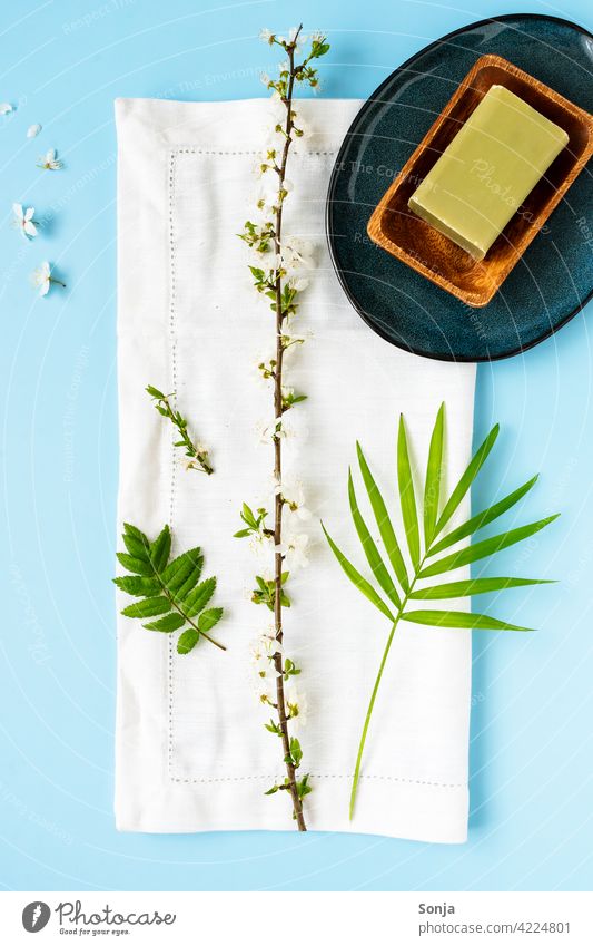 Eine grüne Seife in einer Holzschale, ein weißes Handtuch und ein Blütenzweig auf einem blauen Hintergrund Beautyfotografie Wellness Spa Erholung Hautpflege