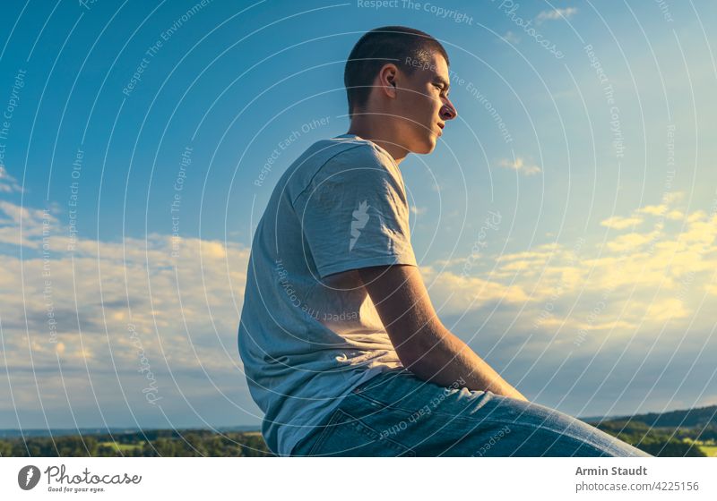 sitzender junger Mann im Profil schaut in den Sonnenuntergang Sitzen Blick Landschaft Himmel blau Sommer im Freien PKW Natur Sonnenlicht Sonnenstrahlen