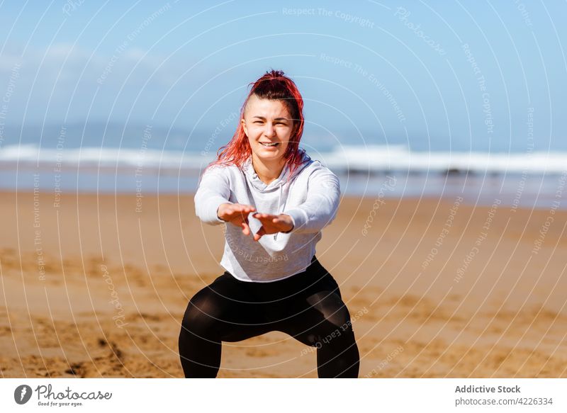 Lächelnde Sportlerin in Kniebeuge-Pose am Meeresufer Übung Training Energie Vitalität Porträt Frau Athlet Gesunder Lebensstil Wellness ausdehnen üben Aufwärmen