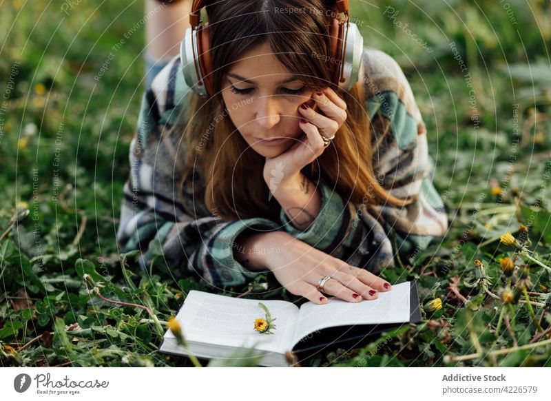 Frau mit Kopfhörern liest ein Buch im Gras lesen Literatur Wissen freie Zeit zuhören benutzend Apparatur Gerät Lehrbuch Bildung Musik Melodie tausendjährig