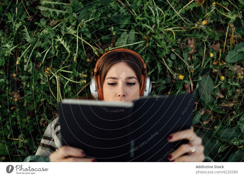 Frau mit Kopfhörern liest ein Buch im Gras lesen Literatur Wissen freie Zeit zuhören benutzend Apparatur Gerät Lehrbuch Bildung Musik Melodie tausendjährig