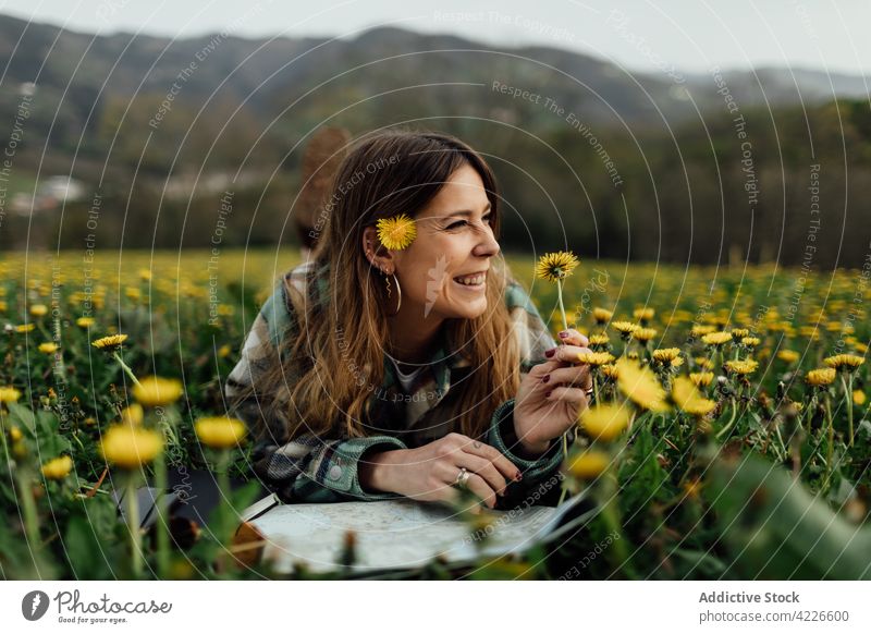 Glücklicher Tourist mit Landkarte, der sich in einem Feld mit blühendem Löwenzahn ausruht Reisender Lächeln Urlaub Blütezeit Blume Landschaft Frau Natur