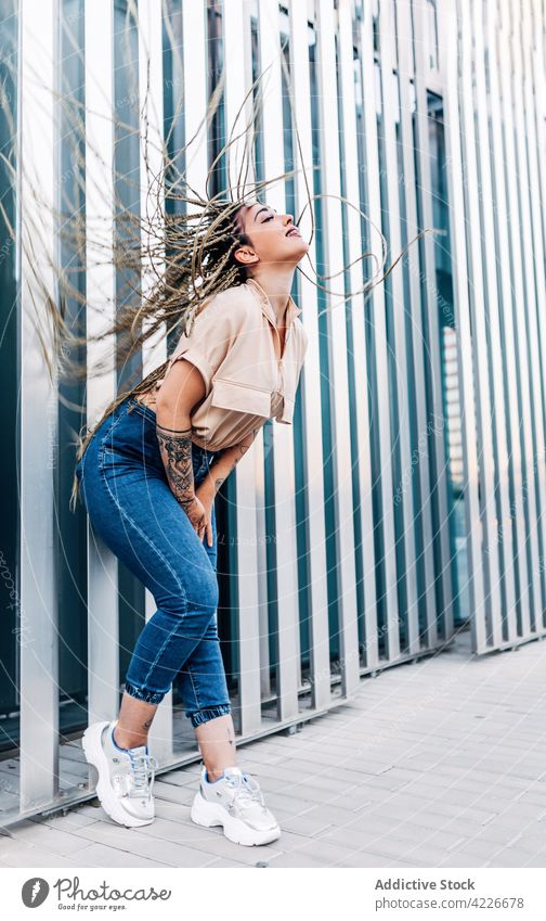Trendige Frau mit fliegendem Haar auf einer Stadtstraße fliegendes Haar Mode Stil Individualität cool Tattoo Geflecht genießen nach vorne lehnen Wand Turnschuh