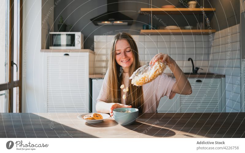 Lächelnde Frau gießt Maisringe in eine Schüssel in der Küche eingießen Müsli Frühstück vorbereiten Keks Biskuit süß gebacken Tisch heimisch heiter Tasche