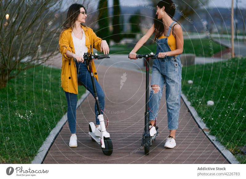 Crop lächelnde Freundinnen mit Roller interagieren auf städtischen Fußweg Tretroller Streetstyle heiter Zeit verbringen Wochenende elektrisch Stadt Rasen Frauen