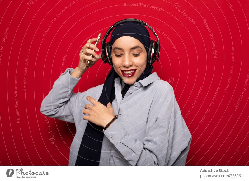 Fröhliche muslimische Frau mit Kopfhörern, die Musik auf ihrem Mobiltelefon hört heiter Tanzen zuhören meloman Glück lässig Gerät Apparatur Drahtlos genießen