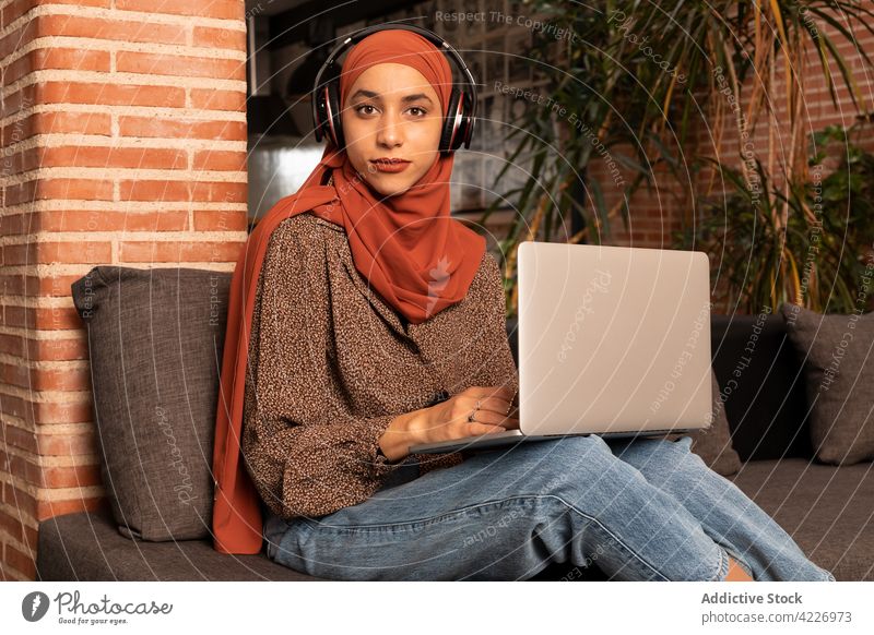 Muslimische Frau arbeitet am Laptop auf dem Sofa Apparatur Kopfhörer benutzend Hijab Surfen Inhalt zuschauen Liege Computer Internet Browsen jung muslimisch