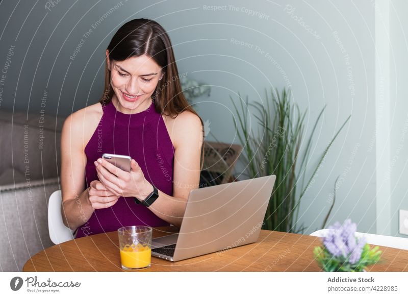 Lächelnde Frau, die ein Smartphone benutzt und mit einem Laptop am Tisch sitzt benutzend Zahnfarbenes Lächeln Apparatur Browsen Telefon Inhalt heiter Netbook