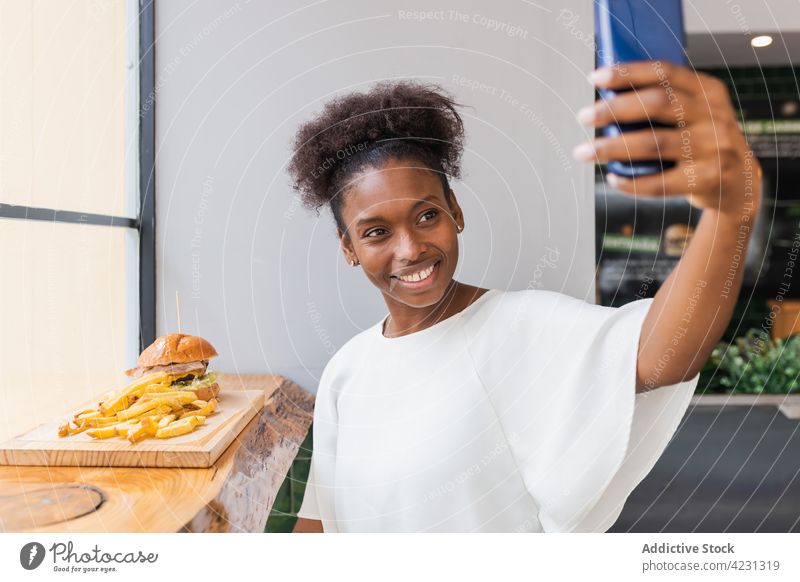 Glückliche schwarze Frau isst Pommes und macht ein Selfie Fries Burger essen freudig Smartphone Restaurant heiter fotografieren Biss lecker Mittagessen Gerät