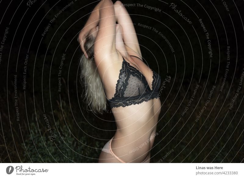 Ein wunderschönes blondes Dessous-Modell streckt sich sinnlich. Ihre sexy Kurven sind überall auf dem Bild zu sehen. Schwarze Bralette und rosa Höschen, das ist alles, was sie in der Nacht trägt. Der dunkle Hintergrund kontrastiert perfekt mit dem Licht auf ihrem Körper.