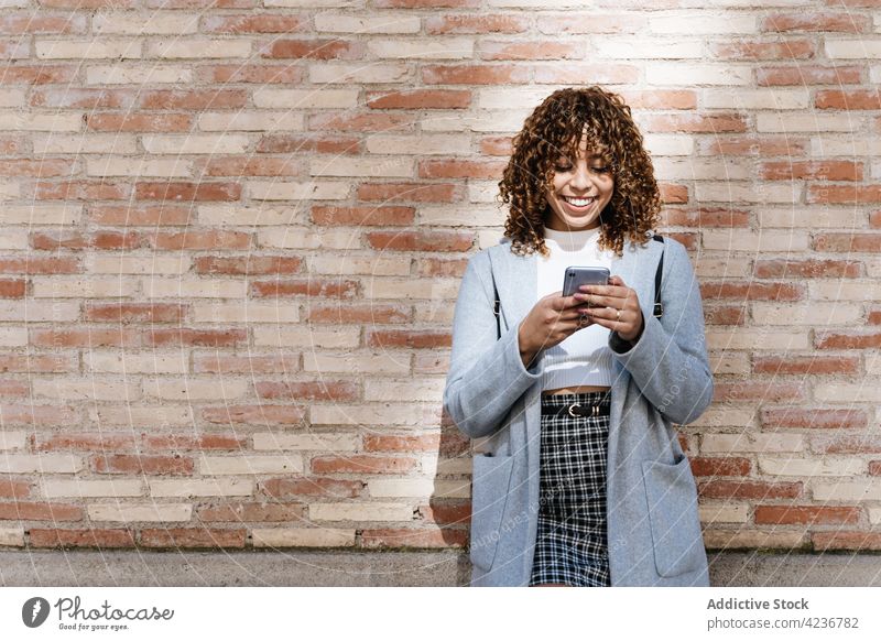 Fröhliche ethnische Frau mit Smartphone an einer Backsteinmauer Zahnfarbenes Lächeln Backsteinwand Browsen heiter Kommunizieren Texten Glück soziale Netzwerke