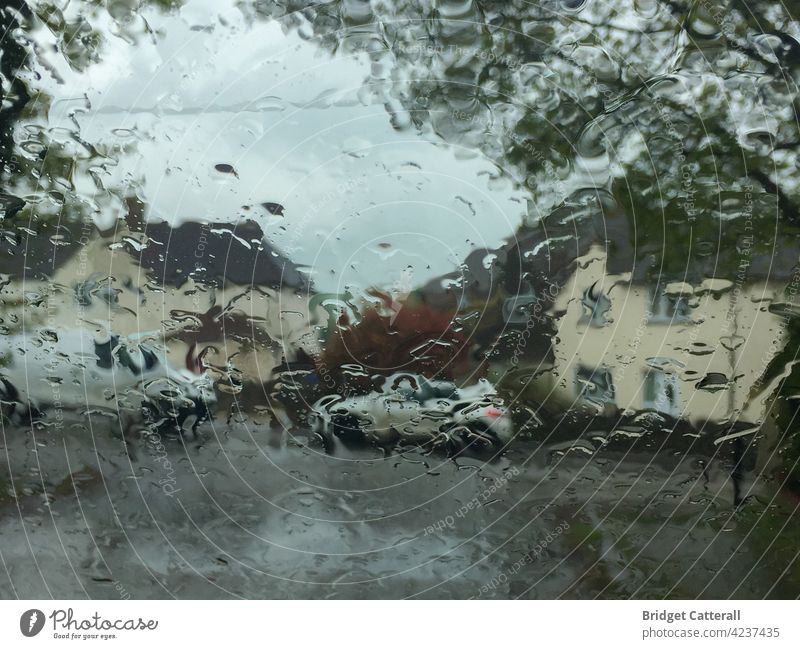 Ein Lieferwagen und ein Auto, die am Straßenrand neben zwei weißen Häusern geparkt sind, aufgenommen durch ein Fenster und gefiltert durch Regentropfen.
