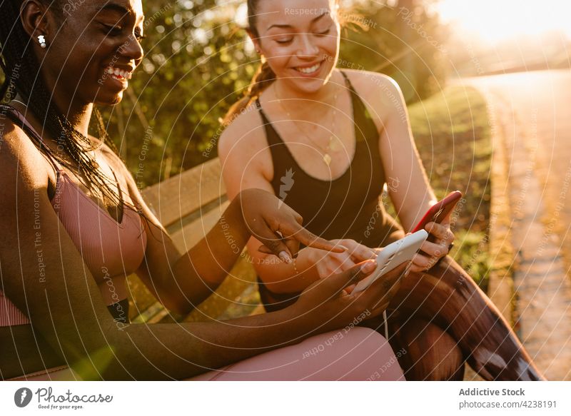 Lächelnde multiethnische Sportlerinnen, die im Park auf ihren Smartphones surfen Browsen Athlet benutzend Surfen Sonnenuntergang Zusammensein Freund Zeigen