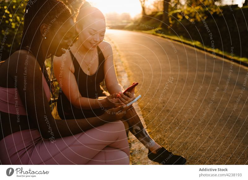 Lächelnde multiethnische Sportlerinnen, die im Park auf ihren Smartphones surfen Browsen Athlet benutzend Surfen Sonnenuntergang Zusammensein Freund Frau
