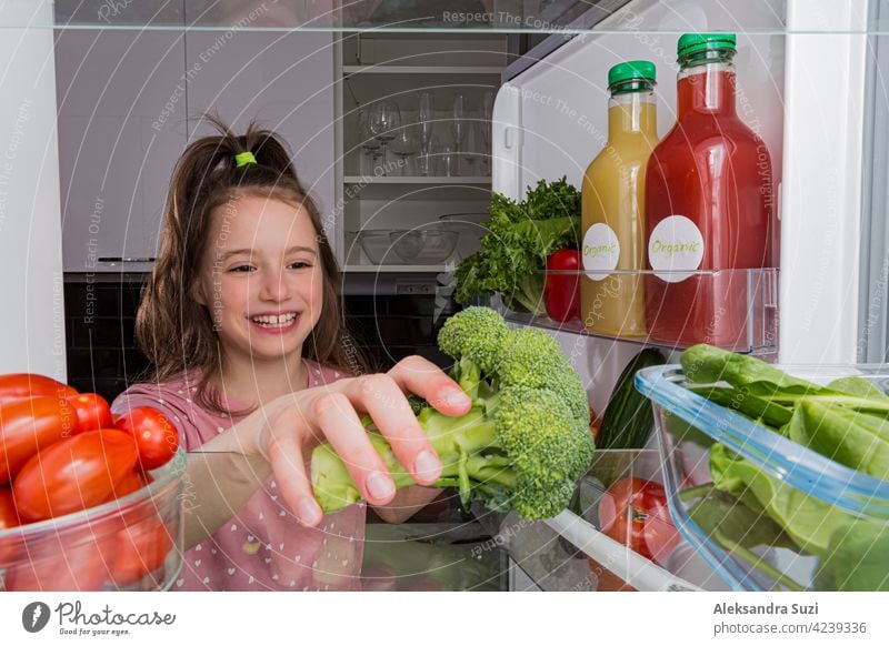 Öffnen Sie den Kühlschrank von innen, Glasregale mit buntem Gemüse, Flaschen mit Bio-Säften. Nettes kleines Mädchen mit glücklichem Lächeln nimmt Brokkoli. Gesunde Ernährung, veganes Konzept.