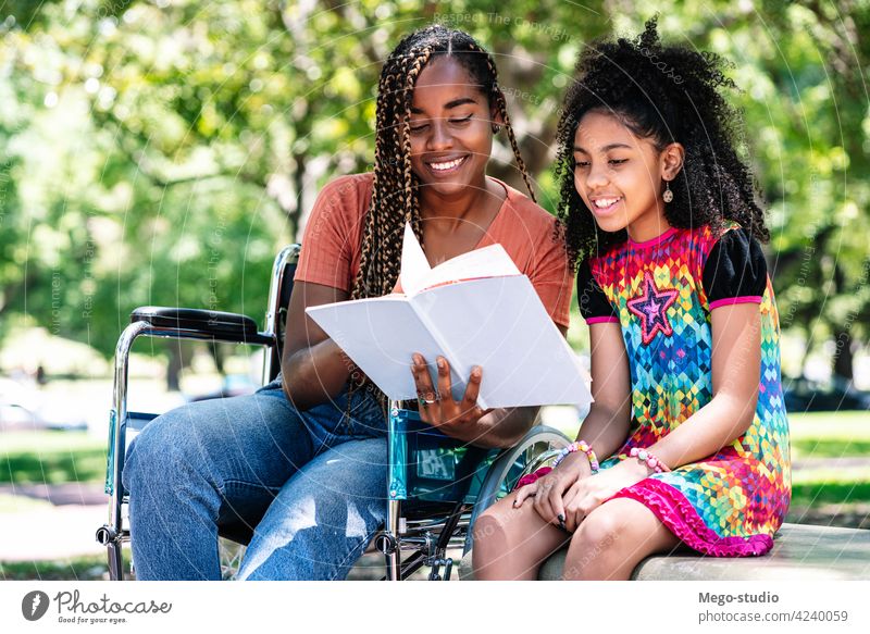 Frau im Rollstuhl liest mit ihrer Tochter ein Buch. Park deaktiviert lesen schwarz Zusammensein im Freien Pflege Lifestyle Tag heiter Freizeit genießend Kind