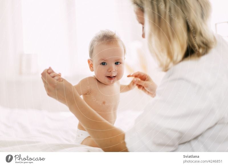Ein süßes Baby sitzt mit ausgestreckten Armen auf dem Bett und hält die Hände seiner Mutter. Neue Fähigkeiten entwickeln, Unterstützung und Pflege Sitzen Kind