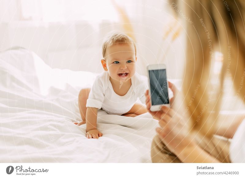 Nettes Baby sitzt auf dem Bett und posiert für seine Mutter, Mutter nimmt Bilder von ihrem Baby auf ihrem Handy. Moderne Technologien, Platz für Text schön Mama